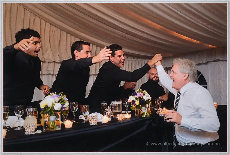 Groomsmen in black celebrate across the head table, trading high fives with a delighted older guest in white.