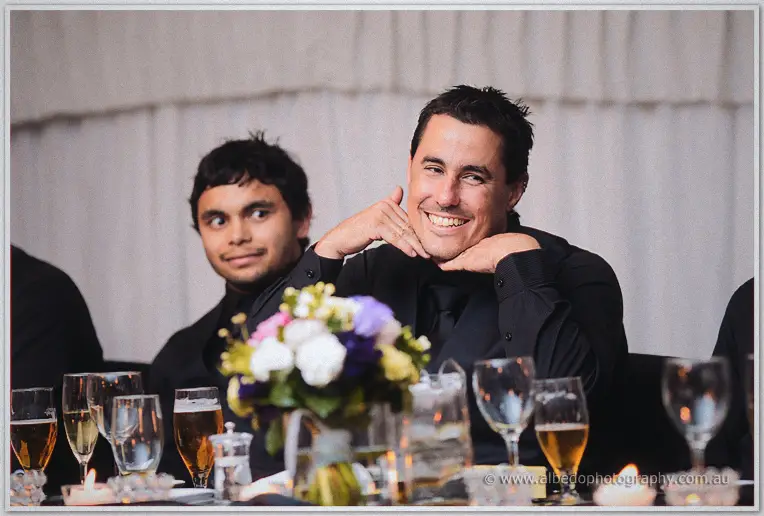 A grinning groomsman rests his chin on both hands at the head table amid wedding flowers and beer glasses.
