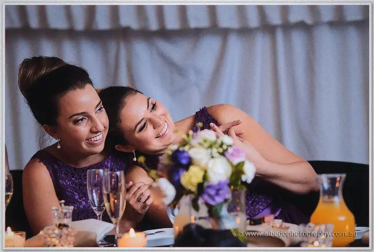 Two bridesmaids in purple lace lean cheek to cheek, smiling at the speeches through the table's flowers.