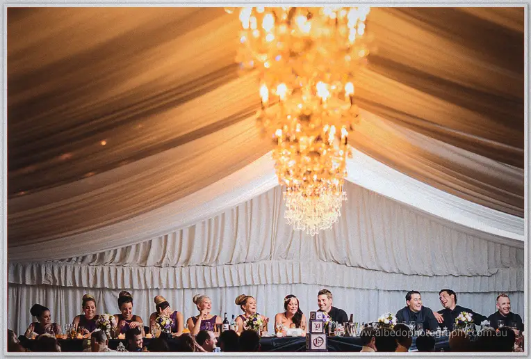 The bridal party and guests sit beneath a glowing crystal chandelier inside the draped white marquee.