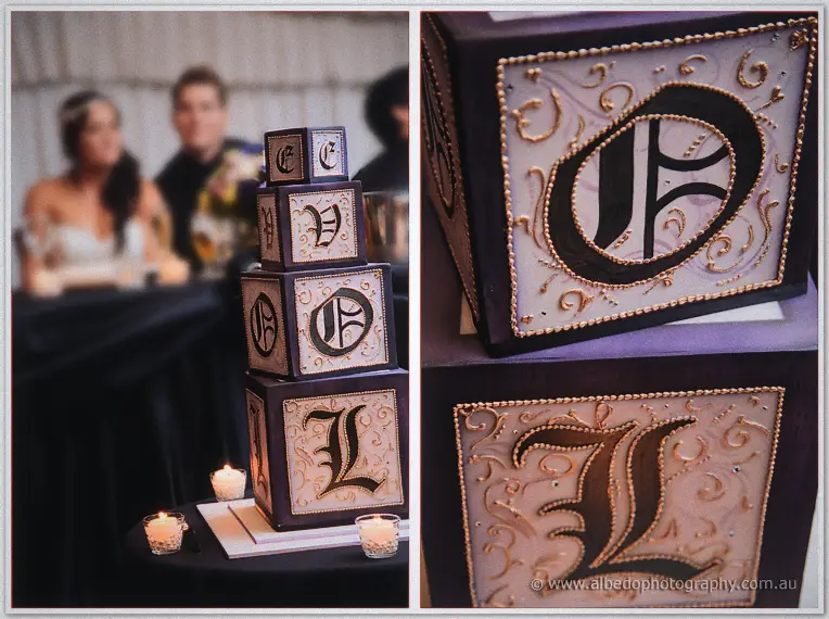 Tiered wedding cake built from lettered boxes with candles, the couple seated behind in the marquee