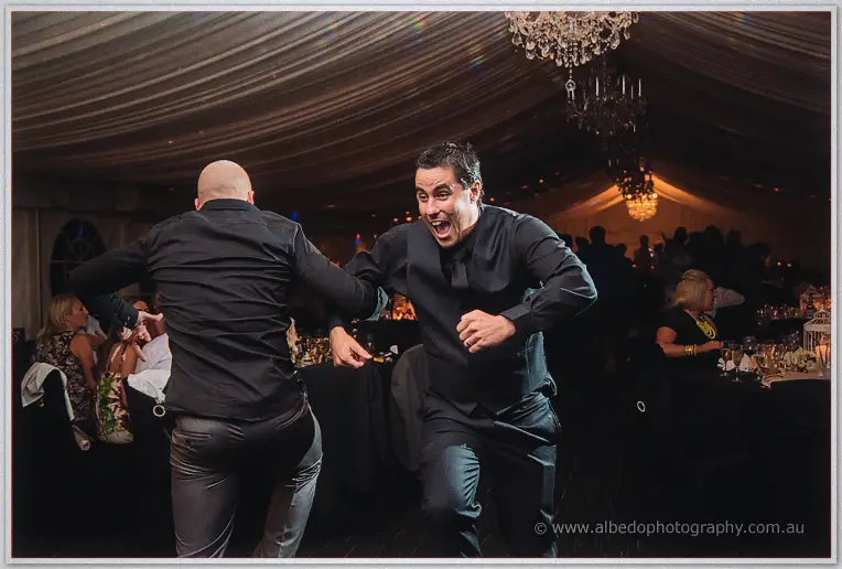 Two groomsmen in black shirts dance hard and laugh under the chandeliers of the reception marquee