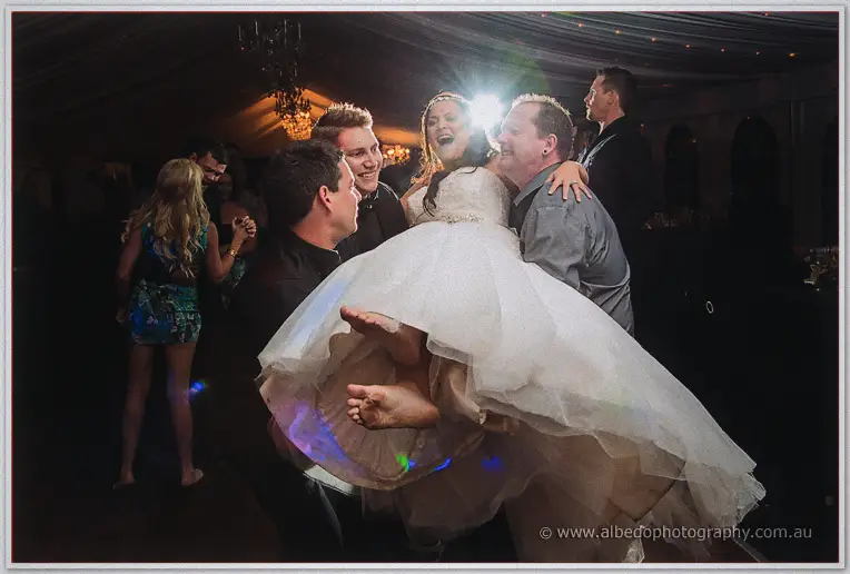 Laughing groomsmen carry the barefoot bride in her tulle gown across the packed dance floor