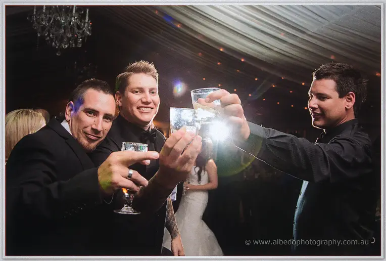 Groomsmen in dark suits raise their glasses in a toast toward the camera amid dancing guests
