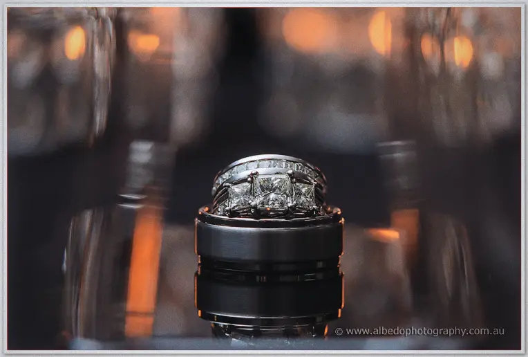 Close-up of Haley's diamond engagement ring stacked on Matt's black wedding band against warm bokeh