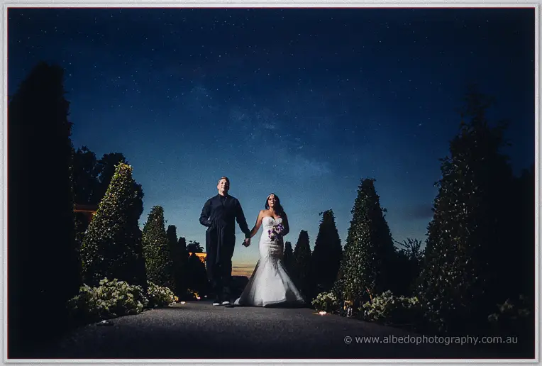 Haley and Matt walk hand in hand down a hedge-lined path under a starry night sky at Caversham House