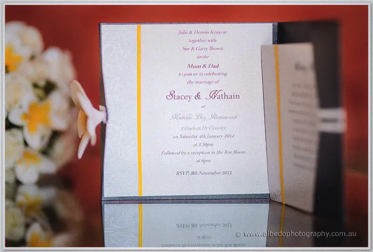 Open invitation card naming Stacey and Nathain propped against frangipanis on a glossy reflective table