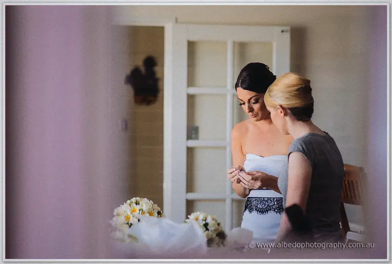 Bridesmaid helping a focused Stacey fasten earrings before a mirror, white and yellow bouquet on the dresser