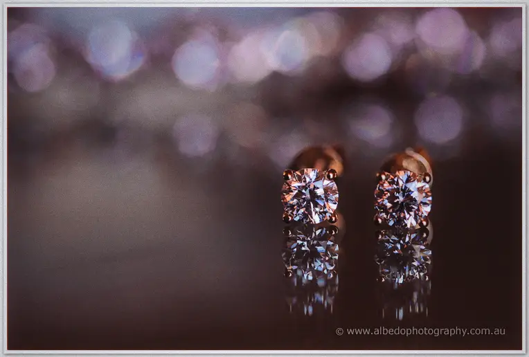 Stacey's diamond stud earrings standing upright on dark glass, sparkling against soft pink bokeh