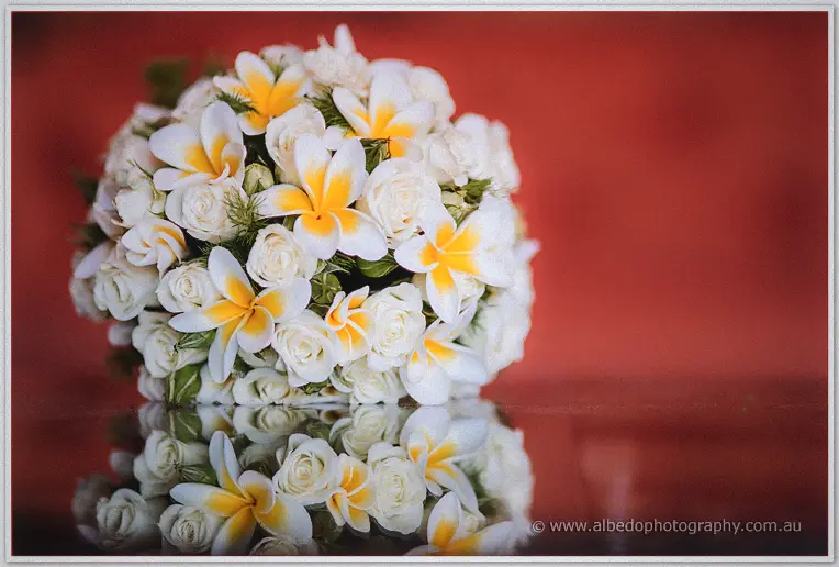 Bridal posy of white roses and yellow-centred frangipani mirrored against a deep red wall