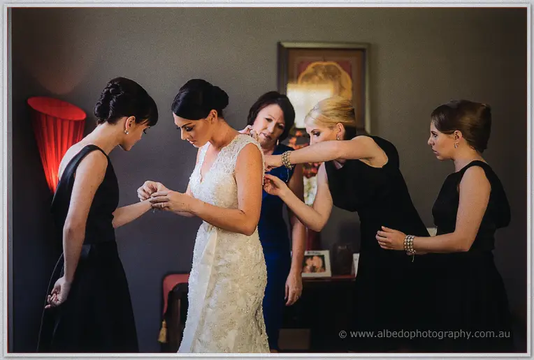 Bridesmaids in black dresses clustering around Stacey to fasten her lace gown as her mother watches