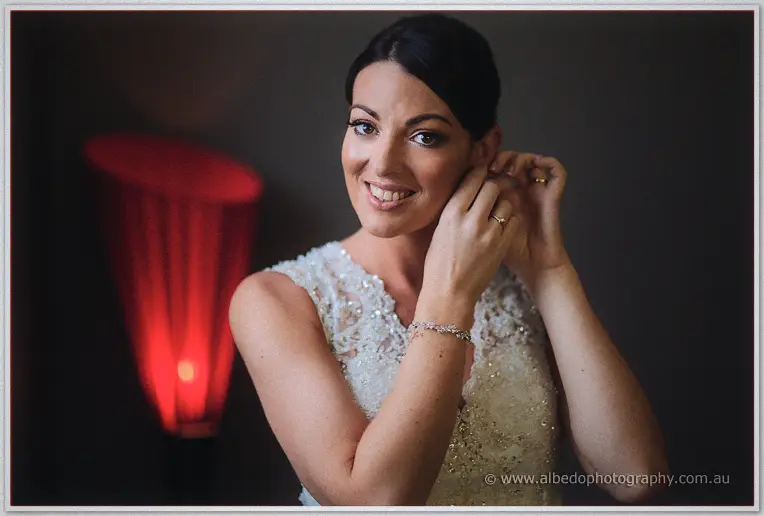 Stacey grinning as she clips in her earring, red lamp glowing beside her beaded lace gown