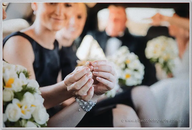 Bridesmaid's hands inspecting the rings in the back seat while bouquets and laughing friends blur around