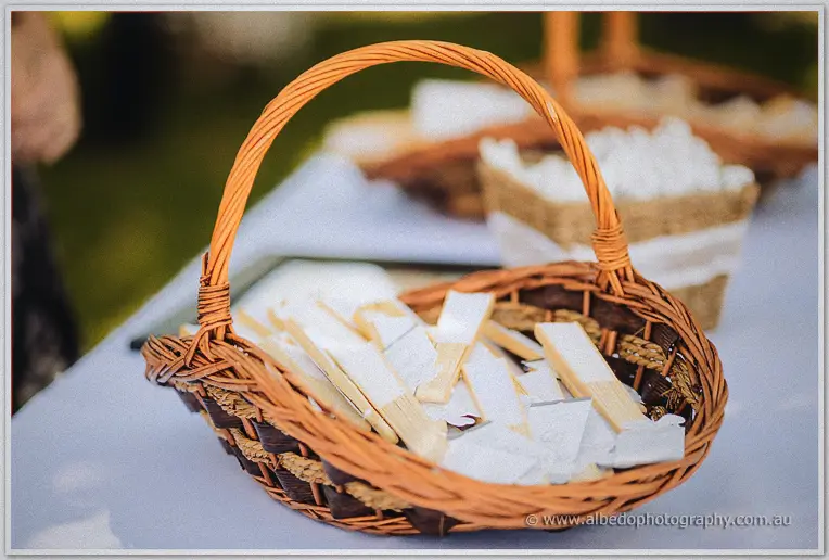 A wicker basket of ribbon-tied ceremony wands rests on a white-clothed table in the sunshine.