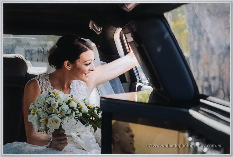 Beaming Stacey waving from the limousine window, frangipani bouquet resting in her lap