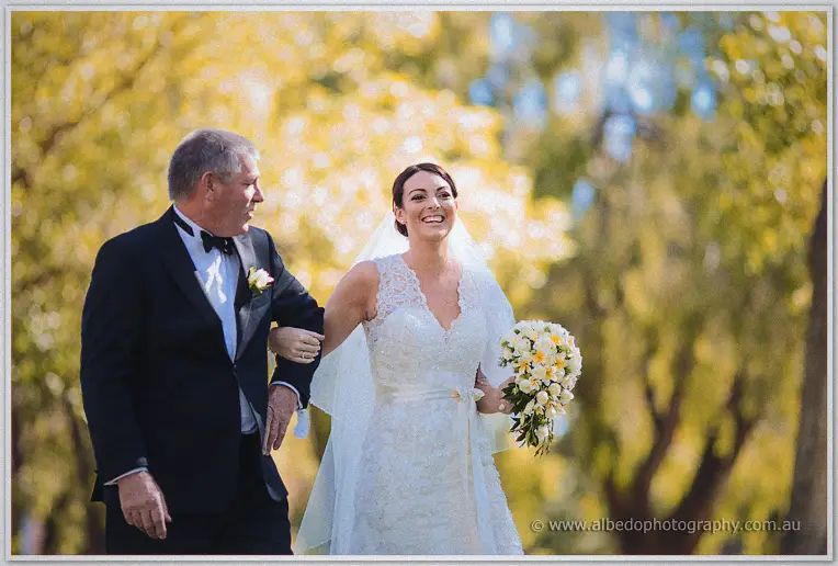 Stacey beams on her father's arm in her lace gown, holding a white and yellow bouquet.