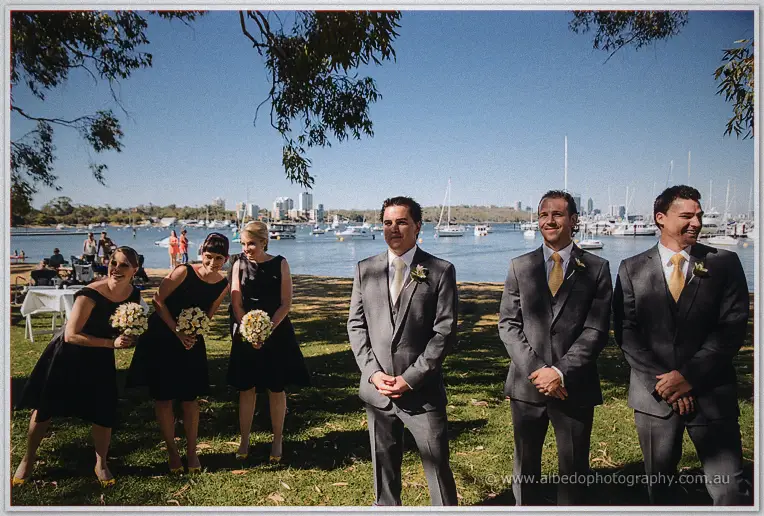 Nathain and his groomsmen wait by the water as bridesmaids in black clutch their bouquets.