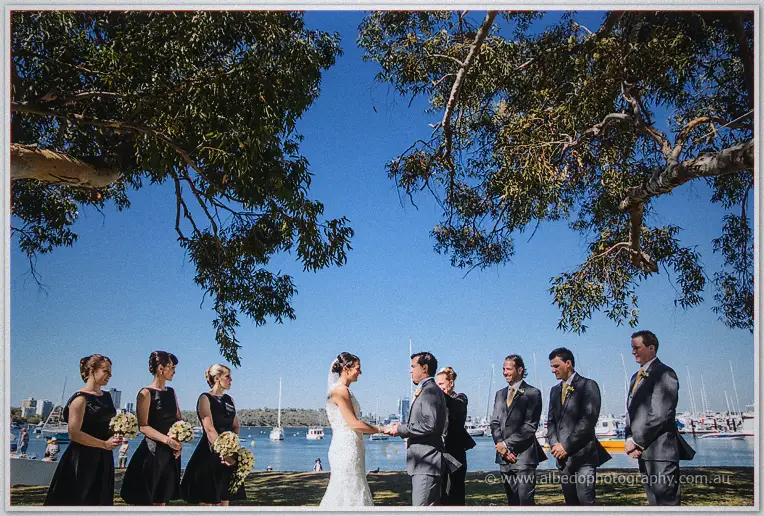 Bridal party flanks Stacey and Nathain as they exchange vows by the sparkling bay.