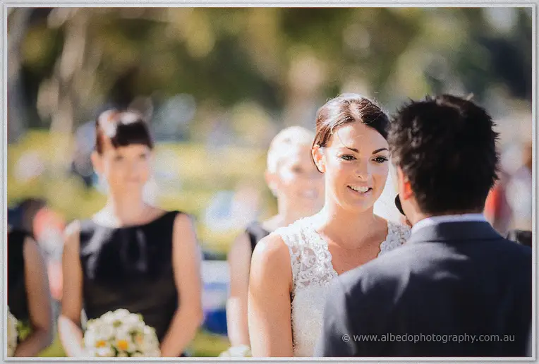 Stacey smiles up at Nathain during their vows, bridesmaids in black softly blurred behind.