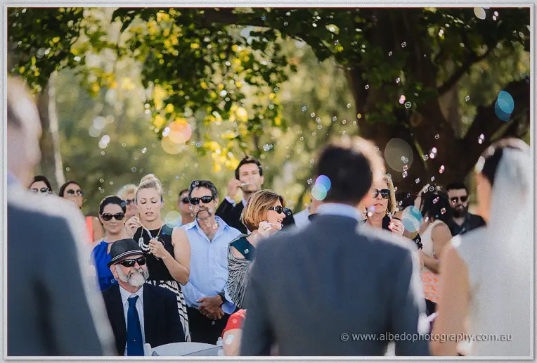 Guests blow soap bubbles over the couple as the sunny ceremony wraps up under the trees.