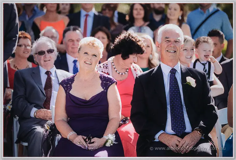 A beaming couple in purple and dark suits watch proudly from the front row.