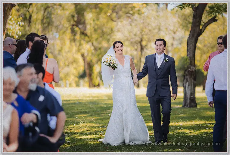 Stacey and Nathain stroll across the lawn hand in hand, bouquet resting on her arm.