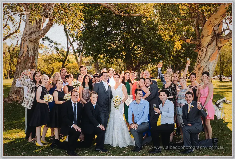 Wedding guests raise champagne flutes around the laughing couple beneath golden-lit gum trees.