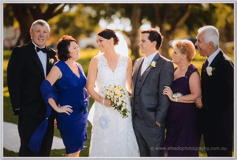 Stacey and Nathain stand arm in arm with their smiling parents in the late-afternoon sun.