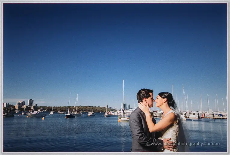 Stacey holds Nathain's face as they kiss before yachts and the city skyline across the bay.