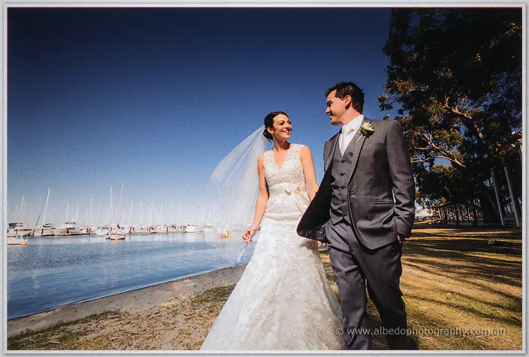The newlyweds laugh together on the sandy shore, veil lifting in the breeze by the moored boats.