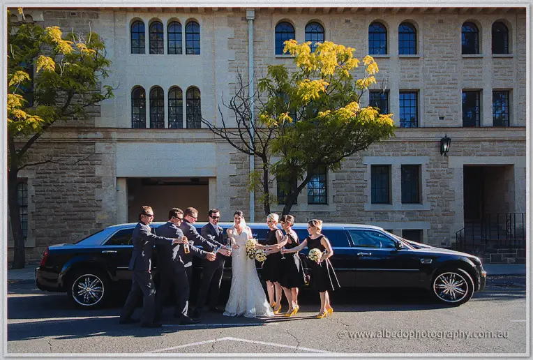The bridal party toasts with champagne beside a black limousine outside a sandstone building.