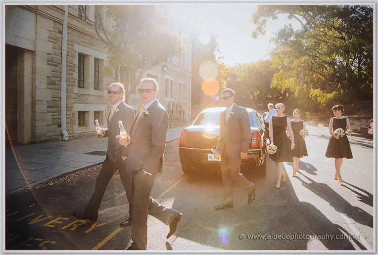 Groomsmen with beer bottles lead bridesmaids across a sun-drenched street in golden flare.