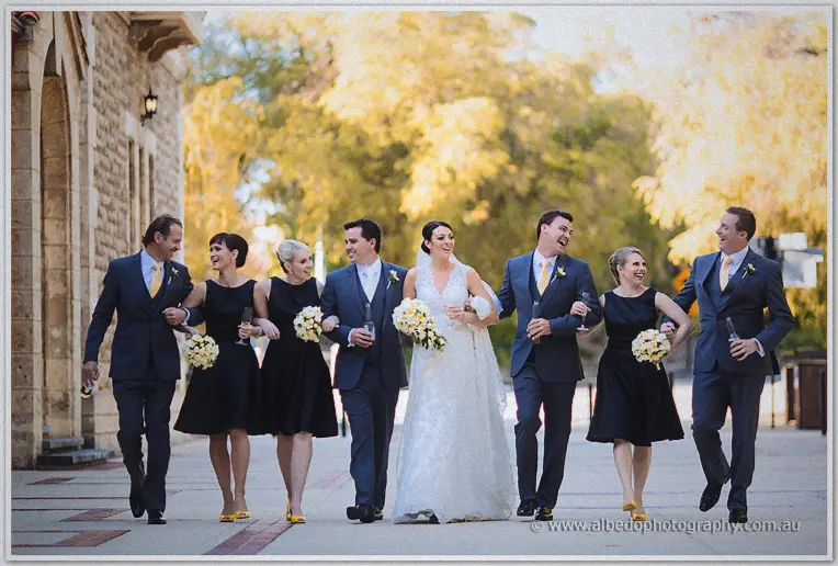 The full bridal party walks arm in arm with champagne flutes past golden trees.