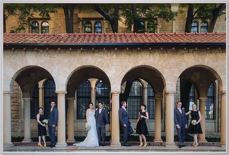 The bridal party poses in the arches of a sandstone colonnade in the warm afternoon light.