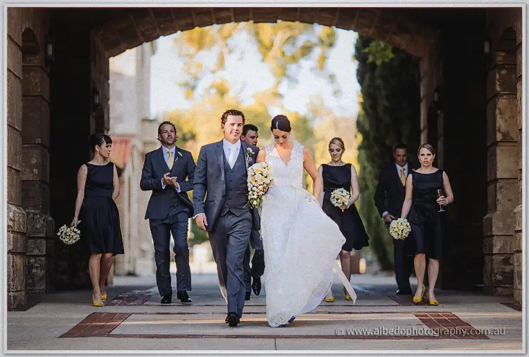 The wedding party strolls through a stone archway, bride and groom leading with bouquets.