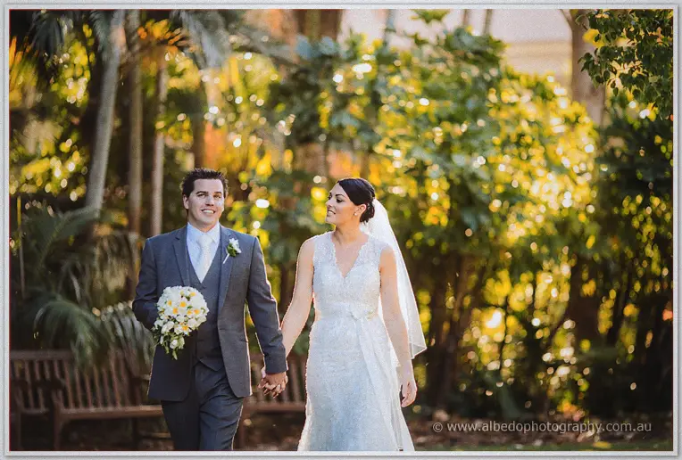 Hand in hand through dappled gardens, Nathain carries the bouquet as Stacey beams at him.