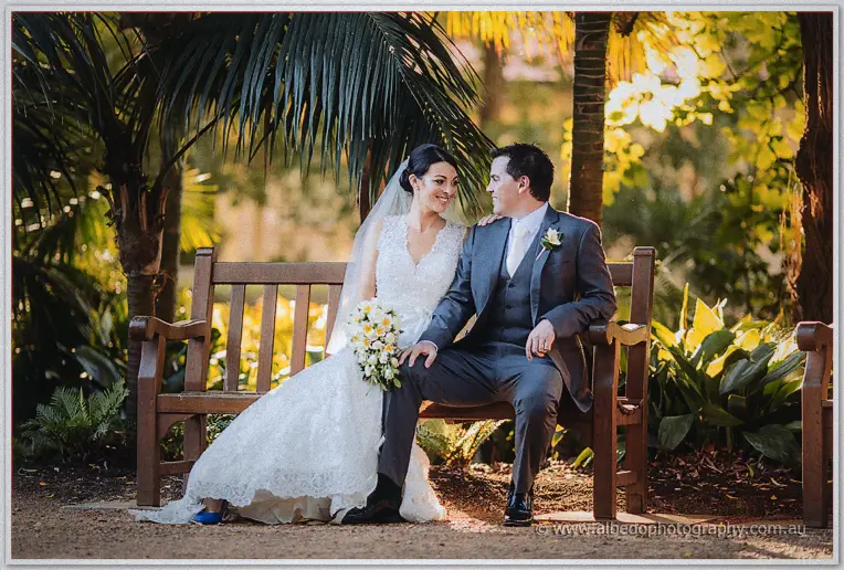 Bride and groom sit close on a timber bench beneath palms, her lace gown pooling in golden light.