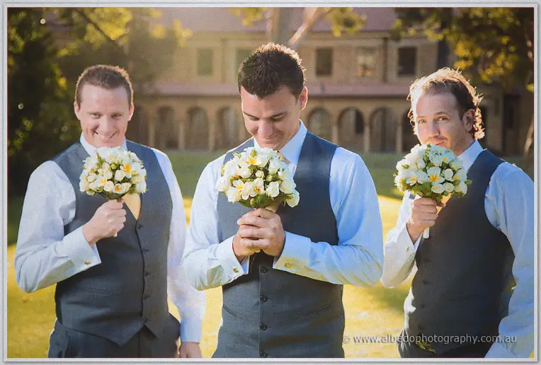 The groom and two groomsmen in grey vests sniff posies on a lawn before a sandstone colonnade.