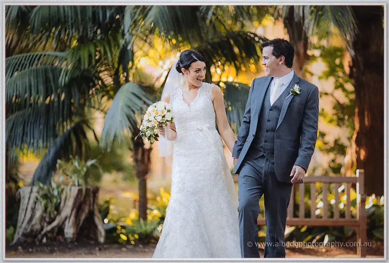Laughing bride and her groom in a grey three-piece suit stroll hand in hand past palms at golden hour.