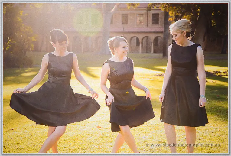 Three bridesmaids in charcoal dresses twirl their skirts on a sunlit lawn, sandstone cloister behind.