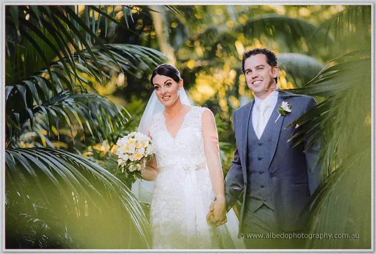 The bride with her white and yellow bouquet and her smiling groom stand framed by lush palm leaves.