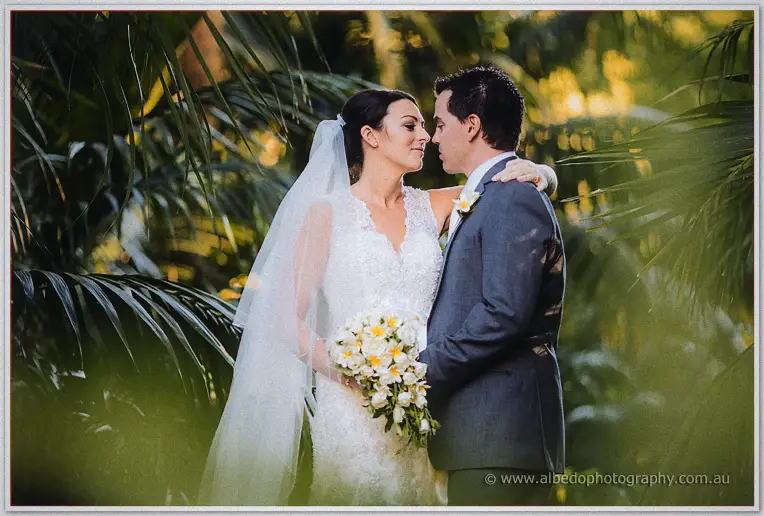 Veiled bride and groom touch foreheads among green fronds, her frangipani bouquet at her waist.