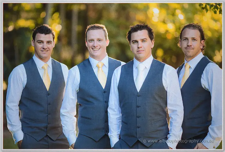 Four groomsmen in grey vests and pale yellow ties line up shoulder to shoulder against golden foliage.