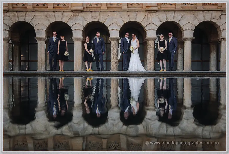 Bridal party pairs line a sandstone arcade, mirrored in the still reflecting pool at their feet.