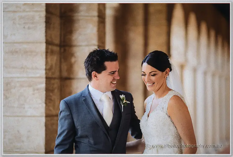 The couple laugh together by a limestone wall, pointed arches falling into soft shadow behind.