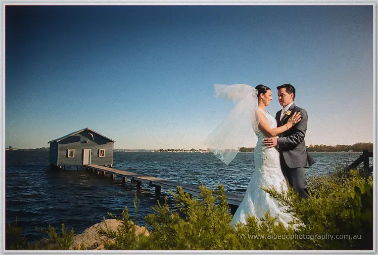 Her veil whips in the river breeze as the couple stand before the blue Crawley Edge boatshed.