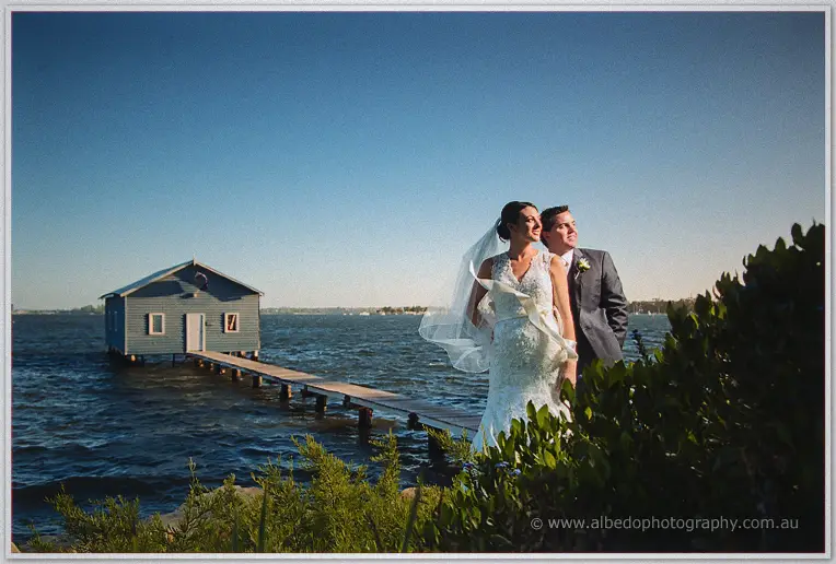 Bride and groom gaze across the Swan River beside the weatherboard boatshed and its wooden jetty.