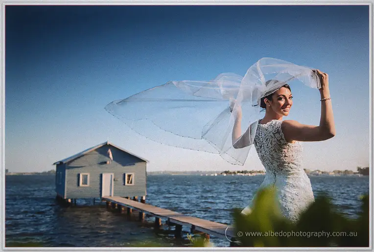 The bride lifts her billowing veil and grins back, boatshed and jetty behind under a deep sky.