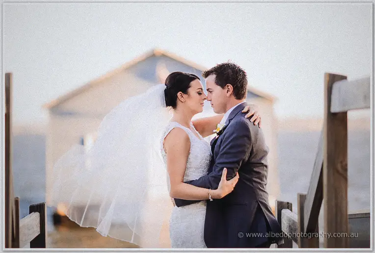 On the jetty the couple press foreheads together, her veil streaming sideways in the wind.
