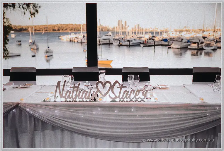 Fairy-lit bridal table with a laser-cut name centrepiece faces the marina, yachts on soft water.